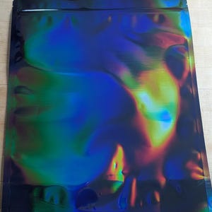 Puede incluir: Una bolsa rectangular iridiscente con borde negro. La superficie de la bolsa refleja un espectro de colores, incluyendo azules, verdes y amarillos. La bolsa está sellada en la parte superior y está plana sobre una superficie de madera clara.