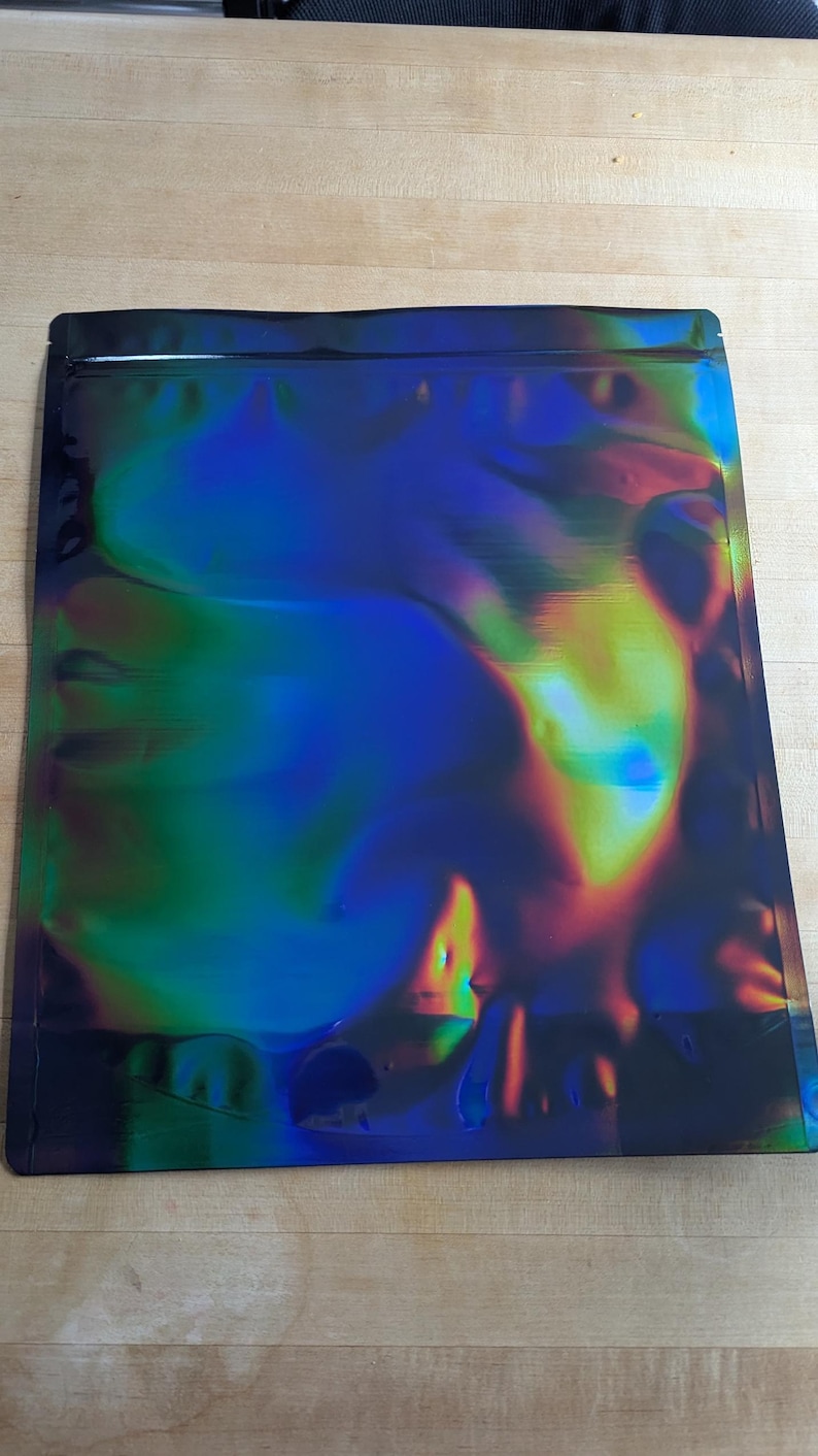 May include: A rectangular, iridescent mylar bag with a black border. The bag's surface reflects a spectrum of colors, including blues, greens, and yellows. The bag is sealed at the top and is laying on a light brown wooden surface.