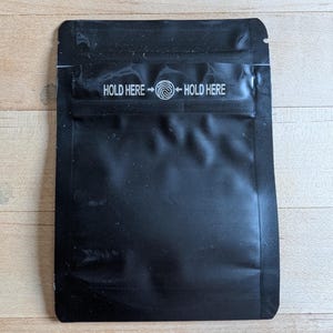 May include: A black, rectangular pouch with a glossy finish. The text "HOLD HERE" is printed in white on the top, with an arrow pointing to a swirl symbol. The pouch is sealed on three sides.