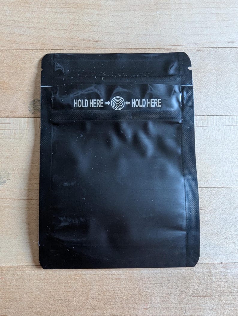 May include: A black, rectangular pouch with a glossy finish. The text "HOLD HERE" is printed in white on the top, with an arrow pointing to a swirl symbol. The pouch is sealed on three sides.