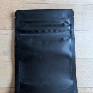 May include: A black, rectangular, sealed pouch with a glossy finish. The pouch has a zip-lock closure at the top and is designed for storing small items. The material appears to be a durable, flexible plastic.