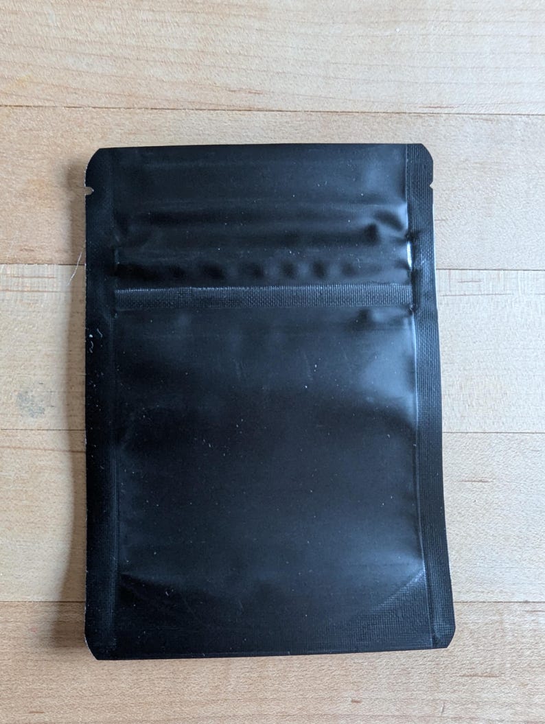 May include: A black, rectangular, sealed pouch with a glossy finish. The pouch has a zip-lock closure at the top and is designed for storing small items. The material appears to be a durable, flexible plastic.