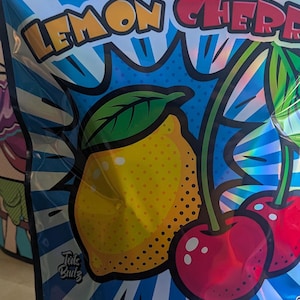May include: A blue and silver package with the words "LEMON CHERRY" in yellow and red. The package features a cartoon illustration of a yellow lemon and two red cherries. The text "Tuts Bulz" is also visible.