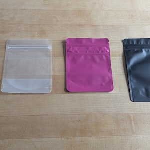 Puede incluir: Cuatro bolsas de pie en fila, cada una de un color diferente. De izquierda a derecha, las bolsas son negras, transparentes, magenta y grises. Cada bolsa tiene un cierre de cremallera y forma rectangular.