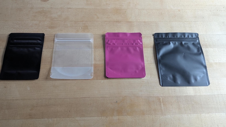 May include: Four stand-up pouches in a row, each with a different color. From left to right, the pouches are black, clear, magenta, and gray. The pouches are rectangular with a zip-top closure.