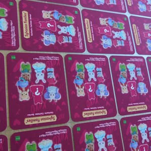May include: Multiple rectangular cards with a magenta background are arranged on a wooden surface. Each card features illustrations of animal characters in various outfits, with the text "Sylvanian Families" and "Natinats Custome Series".