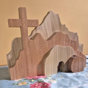 Puede incluir: Escultura de madera de Pascua de una cruz y una tumba. La cruz está hecha de madera marrón claro y la tumba está hecha de madera más clara con una abertura redondeada. La escultura está colocada sobre un mantel floral azul y blanco.