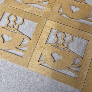 Pode incluir: Conjunto de seis bases de madeira quadradas. Cada base apresenta um design recortado de uma chávena de café com um coração. As bases são feitas de madeira castanha clara e estão dispostas numa superfície cinzenta.