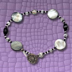 May include: A bracelet with large, round, iridescent beads and smaller black and clear beads. The bracelet has a decorative clasp with an ornate design. The bracelet is displayed on a textured purple surface.