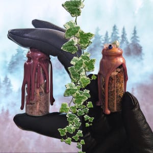 May include: Two small glass bottles filled with herbs and powders, sealed with dripping wax. The bottles are held in a black glove, with a backdrop of a forest scene. Green and white ivy vines are draped between the bottles.