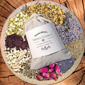 May include: A beige cloth bag labelled "Varanus Witch's Siren's Essence Glamour Spell & Botanical Bath Tea" rests on a ceramic plate. The plate is surrounded by dried flowers and herbs, including rose buds, chamomile, and lavender. The background is a wooden surface.