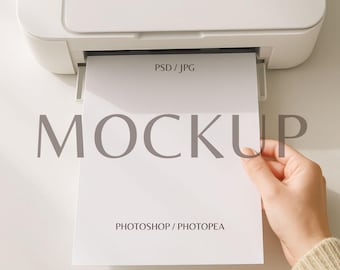Puede incluir: Una impresora blanca con una hoja de papel saliendo de la bandeja de salida. El papel tiene la palabra "MOCKUP" impresa en letras grises grandes, junto con el texto "PSD / JPG" y "PHOTOSHOP / PHOTOPEA".