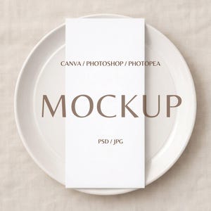 May include: A white rectangular card with the word "MOCKUP" in brown text, placed on a white plate. The card also features the text "CANVA / PHOTOSHOP / PHOTOPEA" and "PSD / JPG". The plate is on a beige surface.