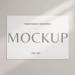 Op de afbeelding: Een witte rechthoekige mockup met het woord 'MOCKUP' in grote, grijze letters. De tekst 'PHOTOSHOP / PHOTOPEA' staat erboven en 'PSD / JPG' eronder. De achtergrond is lichtgrijs met schaduwen.