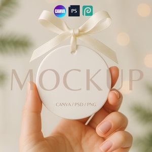 May include: A white, round ornament with a cream-colored ribbon, held by a hand. The ornament has the word "MOCKUP" printed on it, along with the text "CANVA / PSD / PNG". The background is blurred with bokeh lights.