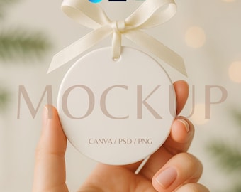 May include: A white, circular ornament with a cream ribbon, held in a hand. The ornament displays the word "MOCKUP" and the text "CANVA / PSD / PNG". The background is blurred with soft, out-of-focus lights.