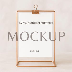 May include: A rose gold metal sign holder with a white paper insert. The paper is held in place by a clip at the top. The word "MOCKUP" is printed in large gray letters on the paper. The text "CANVA / PHOTOSHOP / PHOTOPEA" and "PSD / JPG" are also printed on the paper.