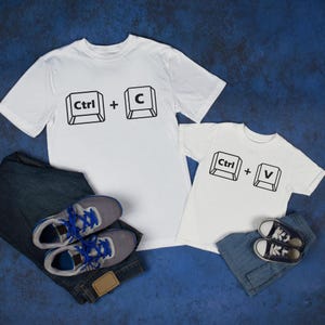 以下が含まれることがあります： 黒いキーボードキーのグラフィックがプリントされた白いTシャツ。1枚のシャツには「Ctrl + C」、もう1枚には「Ctrl + V」と書かれています。シャツは青いジーンズとスニーカーと組み合わせています。コンピューター愛好家向けの楽しいお揃いのセットです。