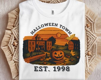 May include: White t-shirt featuring a Halloween-themed graphic. The design includes a pumpkin, buildings, and the text "HALLOWEEN TOWN EST. 1998" in a vintage style. The shirt is folded on a textured surface.