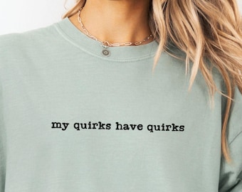 May include: A light green t-shirt with the text "my quirks have quirks" in black. The shirt is made of 100% US cotton and is a relaxed fit. A denim jacket is worn over the shirt.