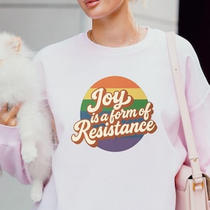 May include: Light pink sweatshirt with the text "Joy is a form of Resistance" in a rainbow circle. The sweatshirt is made of 100% US cotton. A small white dog is held in the image.