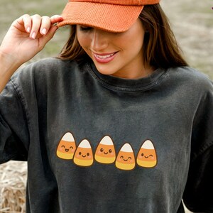 May include: A dark gray t-shirt featuring a row of smiling candy corn graphics. The person in the image is wearing an orange corduroy cap with the text "Pumpkin Spice". Pumpkins and hay bales are in the background.
