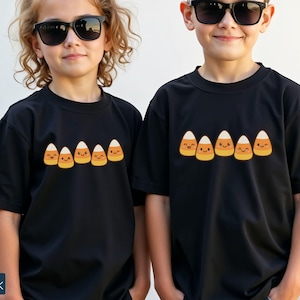 May include: Black t-shirts featuring a candy corn design. The candy corn graphic has a smiling face. The shirts are short-sleeved and worn by two children wearing sunglasses. The word "Black" is visible on the bottom left.