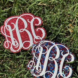 May include: Two decorative acrylic monograms featuring the letters ABC in a white script font. One monogram has a solid red glitter background, while the other features a multi-colored chunky glitter mix of red, blue, and silver flakes.