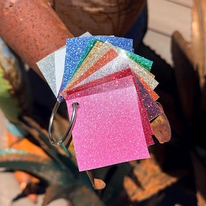 May include: A fan-style swatch set of square glitter material samples held together by a silver metal ring. The collection features various colors including bright pink, red, orange, gold, green, blue, silver, and dark purple, all with a textured finish.