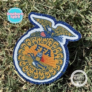May include: A glittery FFA emblem patch featuring a golden eagle, a cross-section of an ear of corn, and a rising sun. The design includes the text Agricultural Education and FFA in blue, with a dark blue glitter border and white outline.