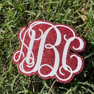 May include: A red glitter monogram with the letters B, C, and B in a decorative script. The letters are white and outlined with a red glitter border. The monogram is set against a background of green grass.