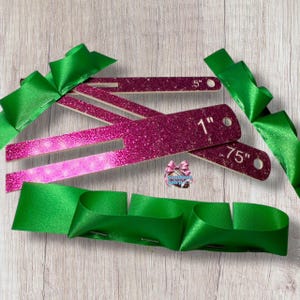 May include: Pink glitter ribbon measuring tools with measurements of 1", .75", and 5" are displayed with green ribbon. The tools are used for measuring ribbon. The tools are pink with white measurements.