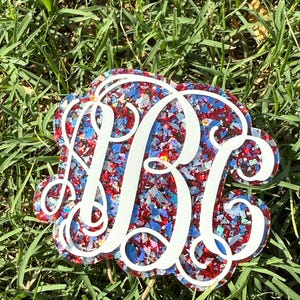May include: A decorative monogram featuring the letters "BKG" in white, layered over a background of red, white, and blue glitter. The design is set against a backdrop of green grass.
