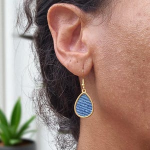 May include: Gold-coloured teardrop earrings featuring a denim blue fabric insert. The earrings hang from a simple hook. The denim fabric has a classic blue jean texture. The earrings are shown on a person's ear.