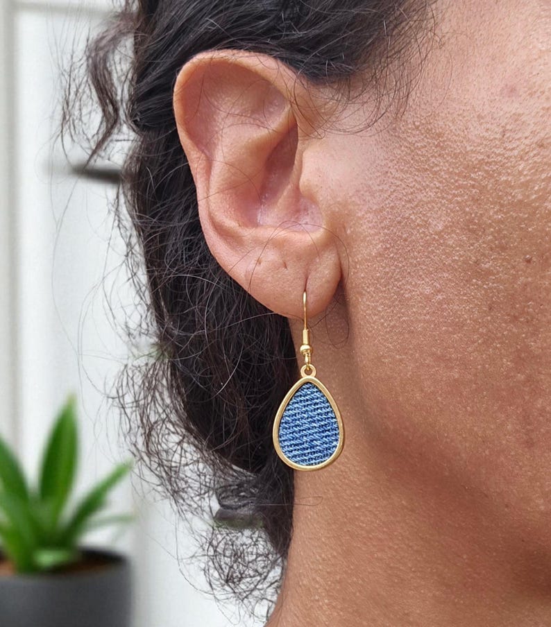 May include: Gold-coloured teardrop earrings featuring a denim blue fabric insert. The earrings hang from a simple hook. The denim fabric has a classic blue jean texture. The earrings are shown on a person's ear.