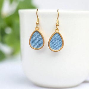 May include: A pair of teardrop earrings with a light blue denim fabric insert framed by a gold-coloured metal border. The earrings hang from gold-coloured ear wires. The background is a white ceramic cup.