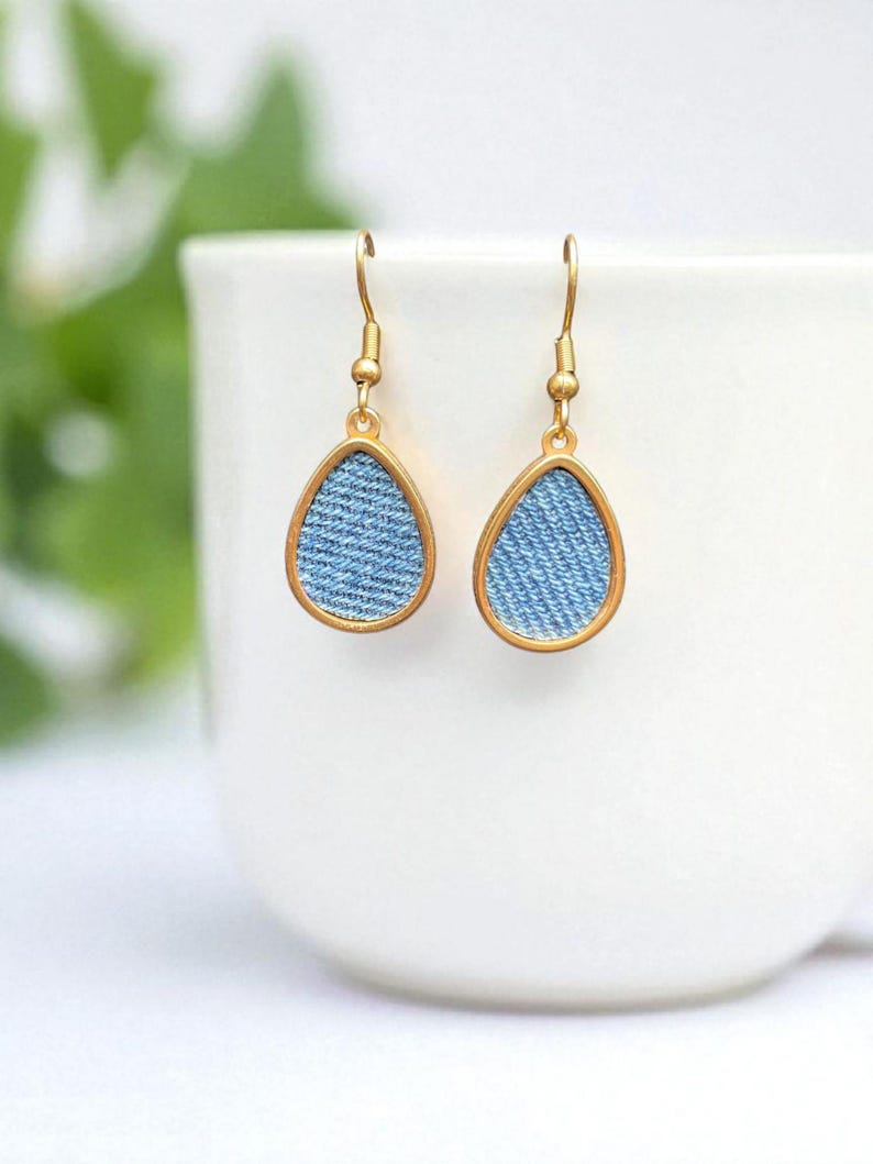 May include: A pair of teardrop earrings with a light blue denim fabric insert framed by a gold-coloured metal border. The earrings hang from gold-coloured ear wires. The background is a white ceramic cup.