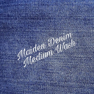 May include: Close-up of blue denim fabric. White cursive text reads "Maiden Denim Medium Wash". The denim has a textured appearance, with a medium blue colour.