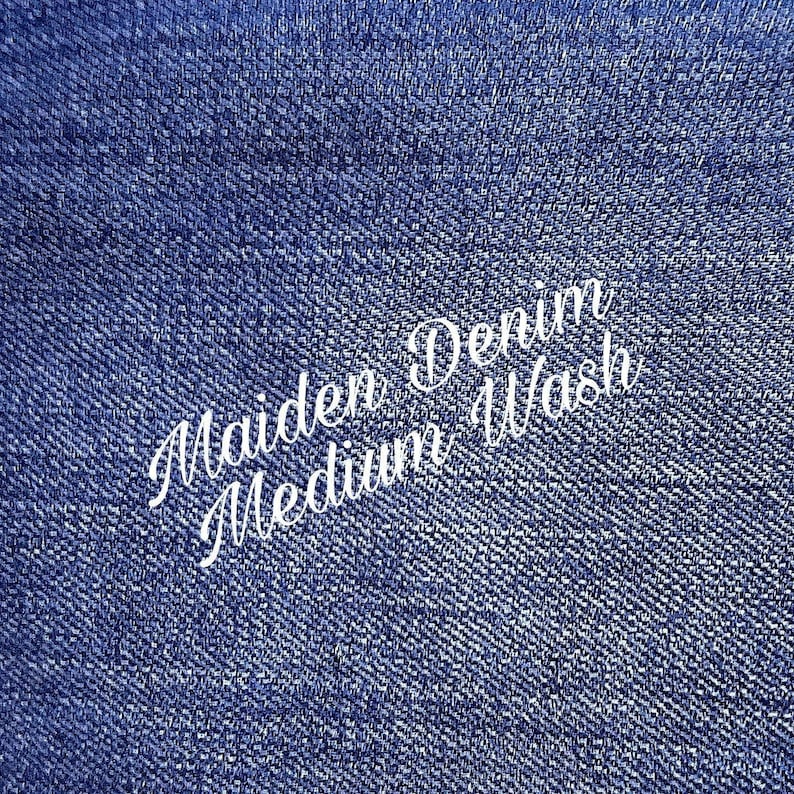 May include: Close-up of blue denim fabric. White cursive text reads "Maiden Denim Medium Wash". The denim has a textured appearance, with a medium blue colour.