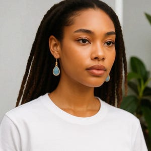 May include: Gold-coloured teardrop earrings with a textured, turquoise-coloured centre. The earrings are hanging from a person's ear. The background is a neutral colour with a green plant.