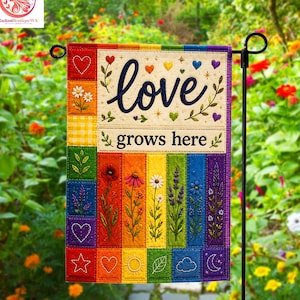 May include: A rectangular garden flag featuring a rainbow-colored vertical panel design. The top section displays the text "love grows here" in dark blue script and print, surrounded by embroidered hearts, flowers, and botanical motifs.