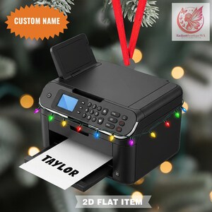 May include: A black printer ornament with a red ribbon for hanging. The printer has a paper with the name "TAYLOR" printed on it. A string of colorful Christmas lights is wrapped around the printer. The ornament is labeled "2D FLAT ITEM".
