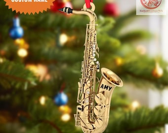 May include: A gold-colored saxophone ornament with the name "AMY" printed on the bell. The ornament hangs from a red ribbon and features the text "CUSTOM NAME" and "2D FLAT ITEM".