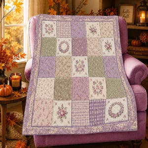 May include: A quilted throw blanket featuring a patchwork design of alternating squares in shades of lavender, sage green, and cream. The fabric squares display various floral patterns, including rose bouquets and wreaths, alongside small checkered prints.