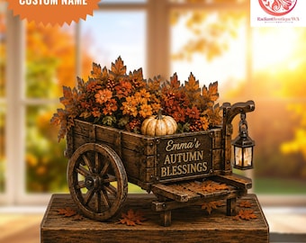 May include: A decorative wooden wagon centerpiece featuring a rustic brown finish, filled with orange and brown autumn foliage and a small white pumpkin. The side of the wagon displays the text Emma's AUTUMN BLESSINGS and includes a hanging lantern.