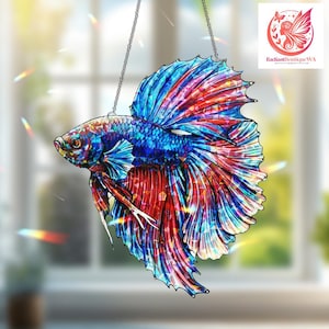 May include: A colorful stained glass betta fish suncatcher. The fish features vibrant blue, red, and purple hues. It hangs from a silver chain. The fish's fins are detailed with intricate patterns. The background is a blurred view of a window.