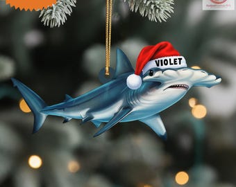May include: A blue hammerhead shark ornament wearing a red Santa hat with the name "VIOLET" on it. The ornament is a 2D flat item and has a gold string for hanging. The background is blurred with Christmas tree branches.