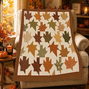 Peut inclure: Une courtepointe rectangulaire présentant un motif répétitif de feuilles d'érable dans des tons d'orange, de brun, de vert olive et de beige. Les feuilles sont sur un fond crème et l'ensemble est bordé d'une bordure en tissu brun uni.