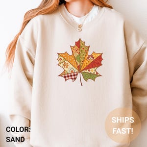 Op de afbeelding: Zandkleurige sweatshirt met een groot, kleurrijk esdoornbladontwerp. Het blad is gesegmenteerd met verschillende patronen in tinten oranje, rood, groen en geel. De tekst "SHIPS FAST!" is rechtsonder zichtbaar.