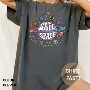 Puede incluir: Camiseta gris oscuro con un diseño de planeta colorido y las palabras "SAFE SPACE". El diseño incluye planetas, estrellas y un anillo alrededor del planeta central. La camiseta es de estilo casual y cómodo.